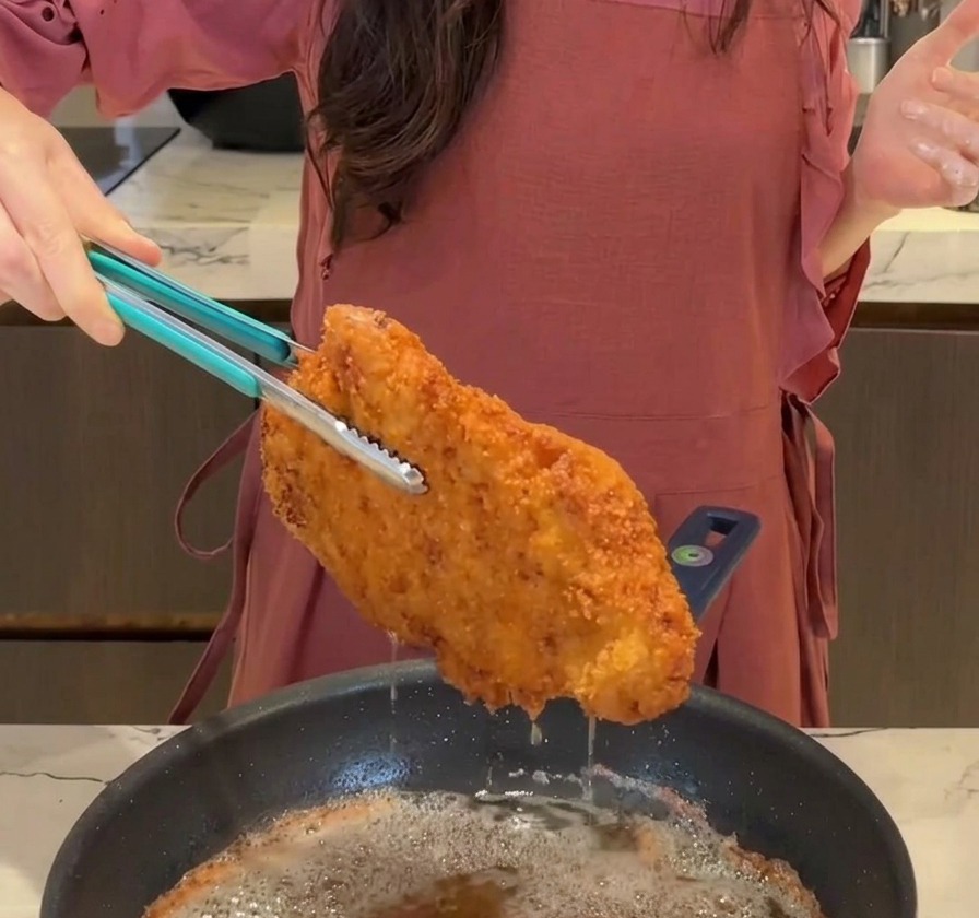 A large, crispy, golden-brown fried pork cutlet being lifted out of a frying pan with blue tongs, with oil dripping back down.