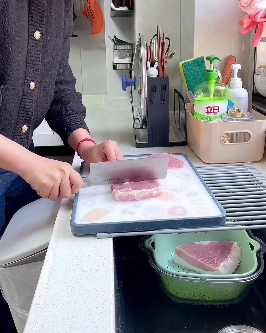 A hand holding a large metal cleaver, using the blunt spine to pound and tenderize a slice of raw pork on a cutting board.