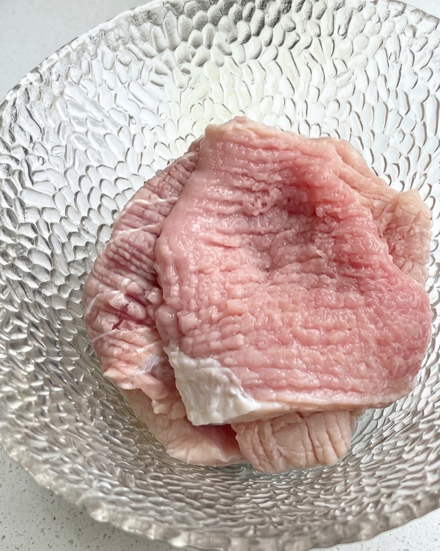 A close-up view of a glass bowl containing raw pork slices topped with chopped green scallions and liquid seasonings.