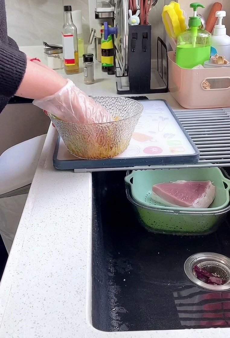 A person wearing a plastic glove mixing thick slices of pork loin with soy sauce, pepper, and starch in a glass bowl.