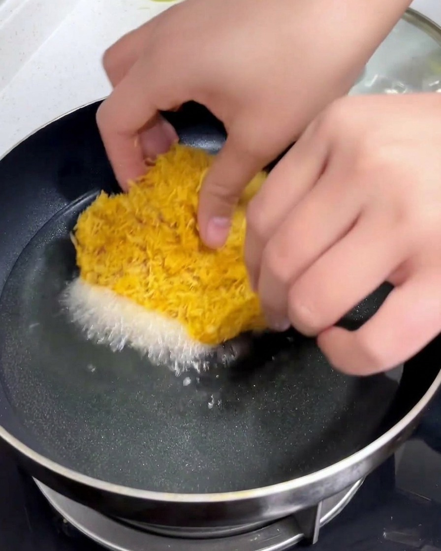 A breaded pork cutlet being lowered by hand into a pan of hot shimmering oil.