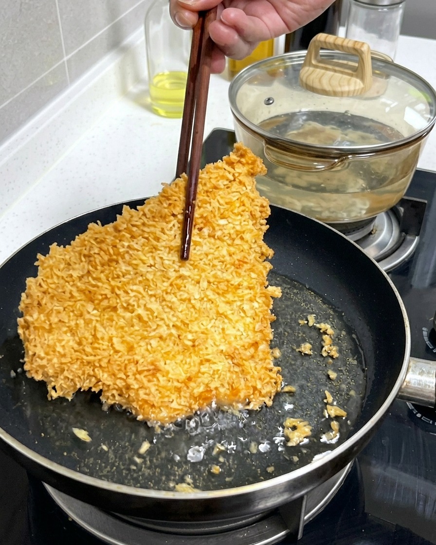 A large, crispy, golden-brown breaded pork cutlet being lifted or flipped with wooden chopsticks in a shallow frying pan with bubbling oil.