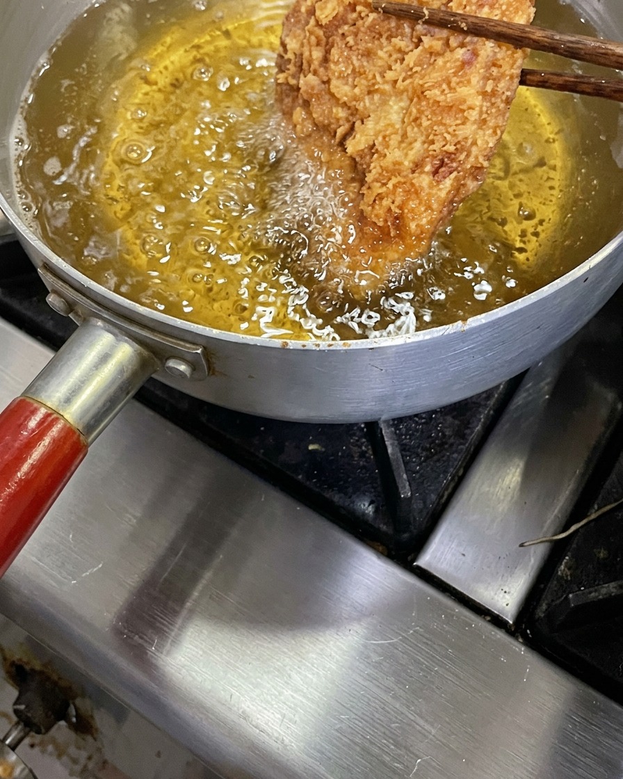 A crispy golden-brown tonkatsu cutlet being lifted with wooden chopsticks from a pot of bubbling hot frying oil.
