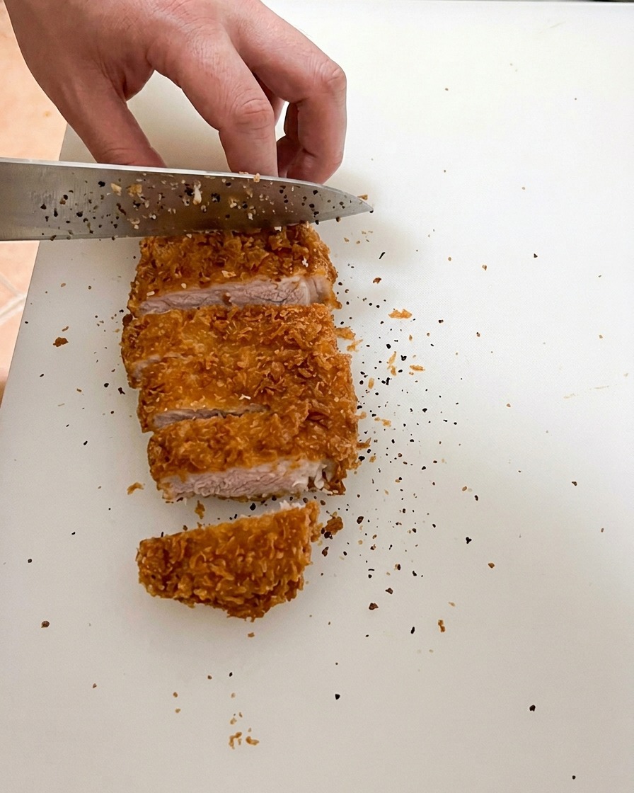 A sharp knife slicing through a perfectly fried pork cutlet on a white cutting board, showing the juicy meat with a light pink interior color.
