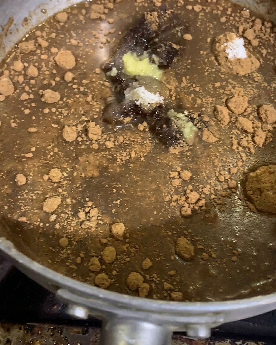 Piles of ground spices, seasoning powders, garlic, and ginger paste sitting atop a thick miso sauce base in a silver saucepan before cooking.