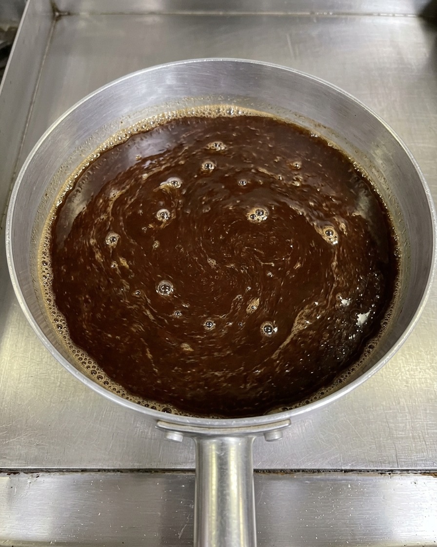 A top-down view of a dark brown miso sauce mixture being stirred with a pair of chopsticks in a small aluminum saucepan.