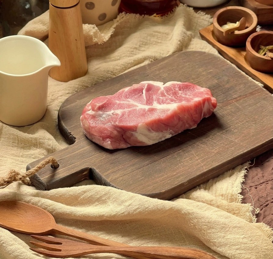 Raw plum blossom pork chop being prepared on a wooden cutting board with kitchen utensils in the background.