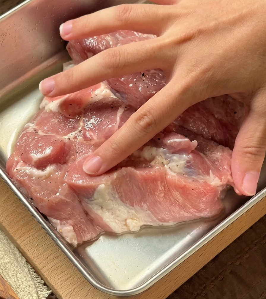 A hand kneading a clear aromatic liquid into raw pork chops inside a metal preparation container.