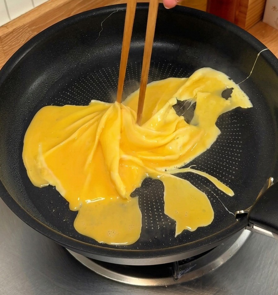 Chopsticks swirling a partially cooked yellow omelet in a black non-stick pan to create a spiral pattern.