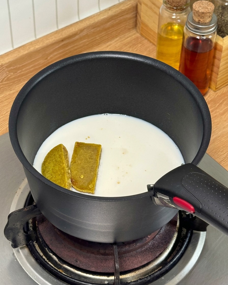 Curry cubes and milk being heated in a small black saucepan on a gas stove.