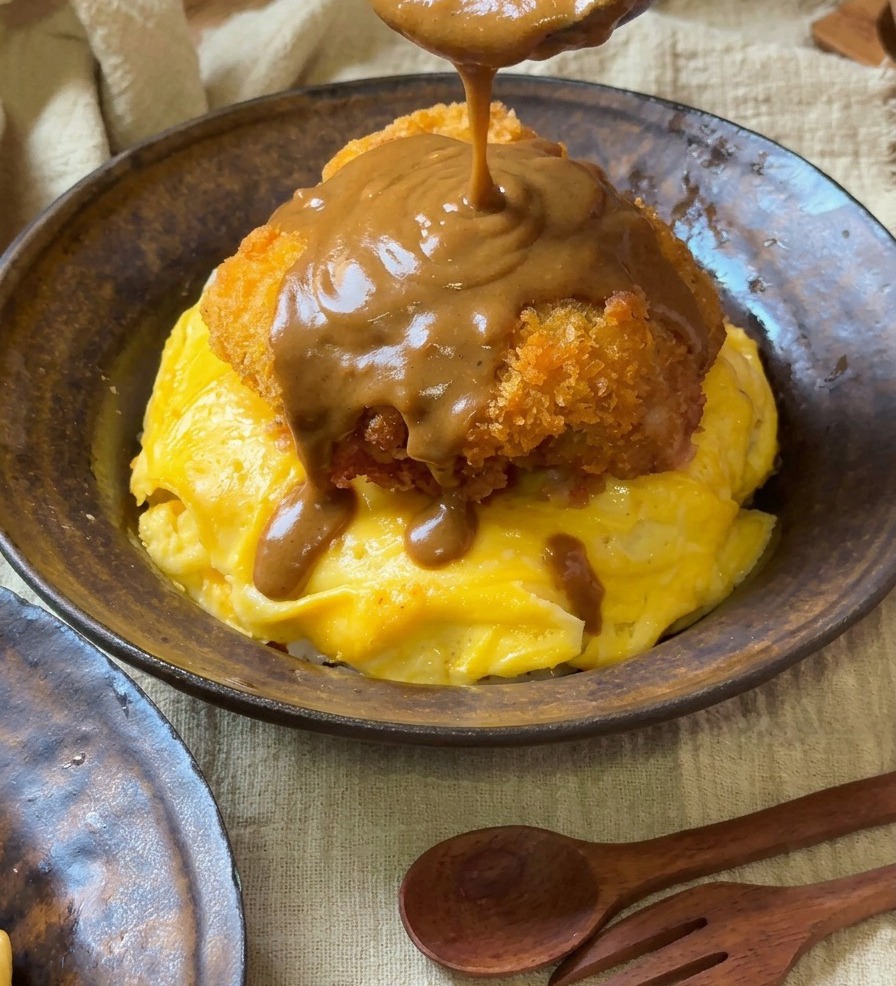 A thick, smooth brown curry sauce being poured from a wooden spoon onto a golden-brown breaded pork chop resting on a bed of swirled yellow eggs.