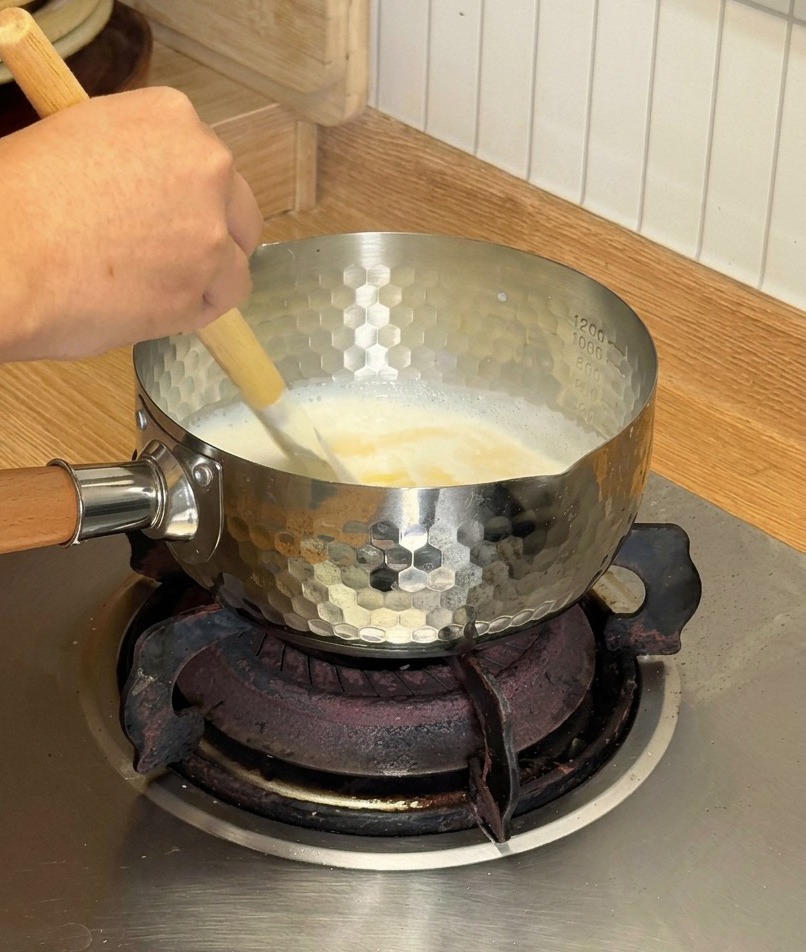 A wooden spatula stirring a creamy white mixture of melting cheese and coconut milk in a small silver pot on a gas stove.