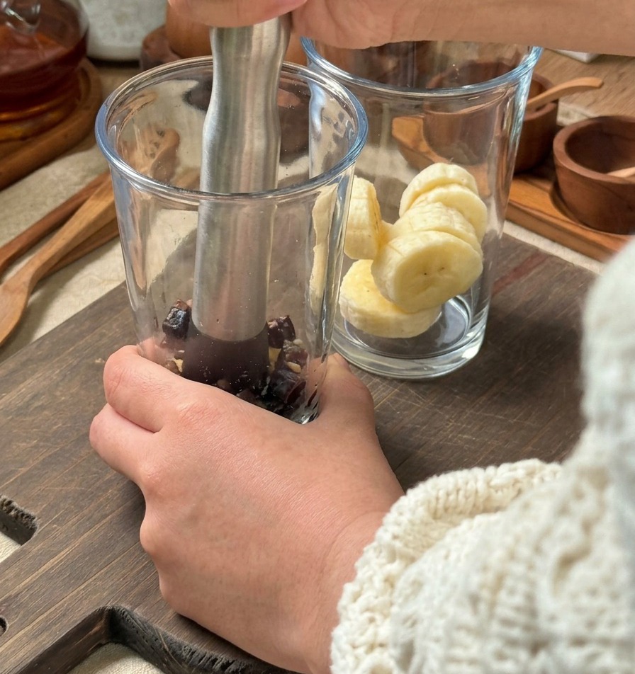 Hands using a silver metal muddler to crush ingredients in a tall glass, with a second glass of sliced bananas waiting nearby on a wooden board.