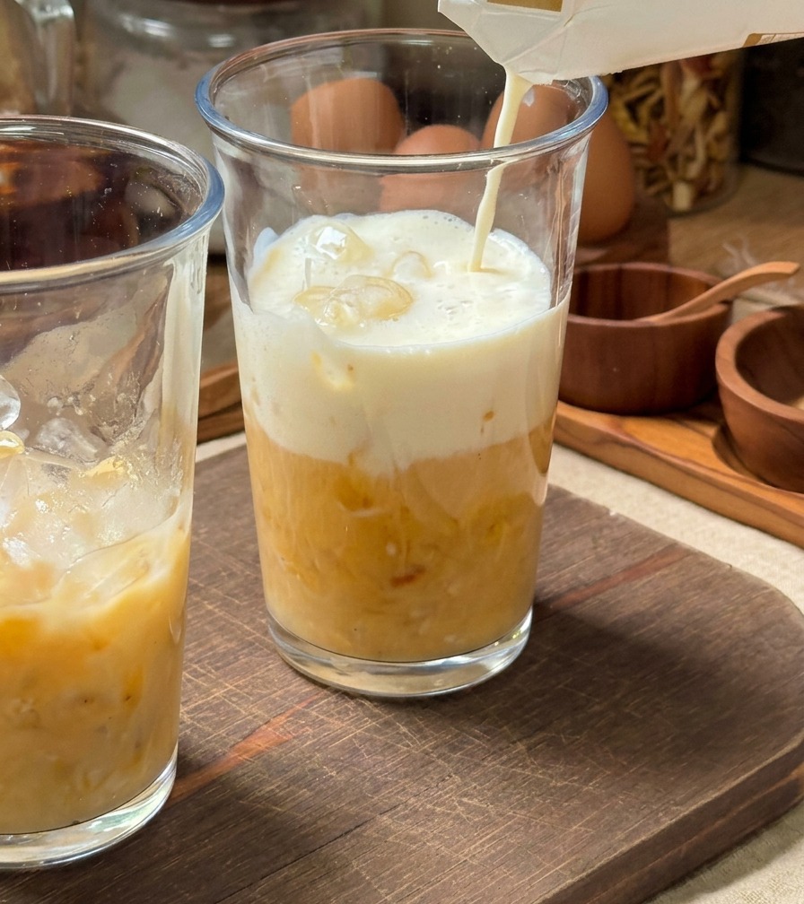 Cold milk being poured from a carton into a tall glass filled with ice and a yellow banana puree base.