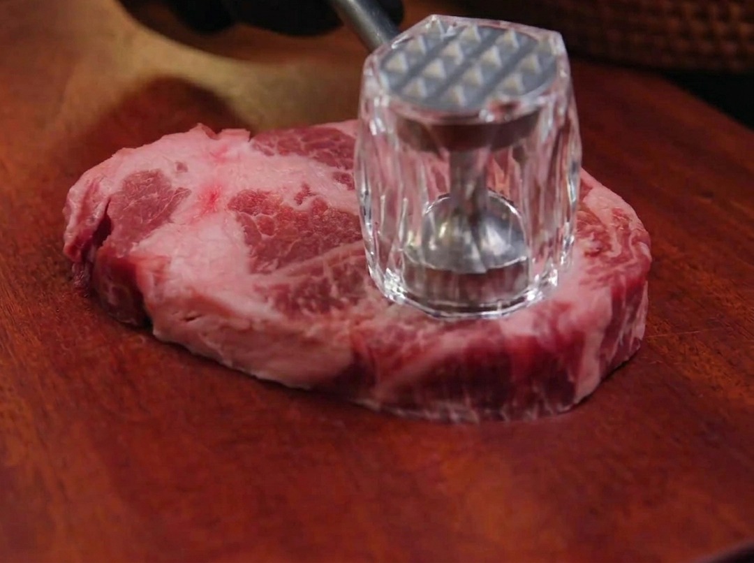 A hand using a metal meat mallet to pound a raw piece of marbled pork shoulder on a dark wooden board.
