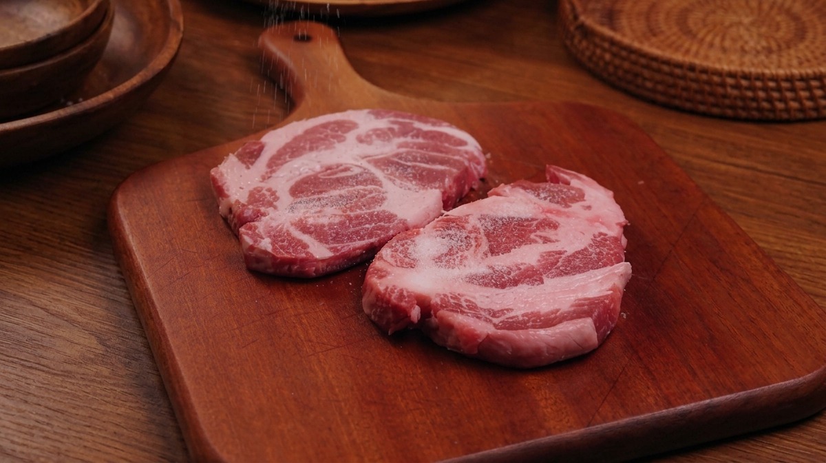 A gloved hand sprinkling salt and pepper over two raw pork steaks resting on a wooden board.