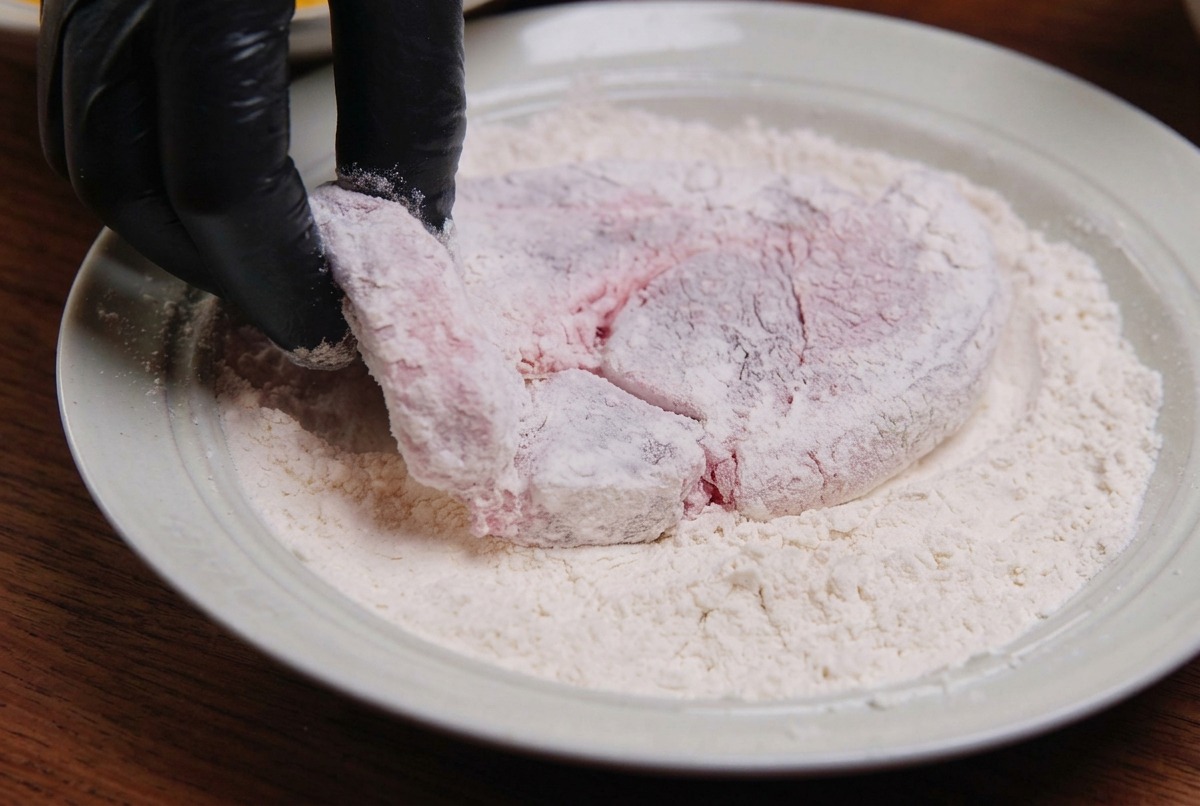 A piece of raw pork being coated in white flour in a white ceramic dish by a gloved hand.