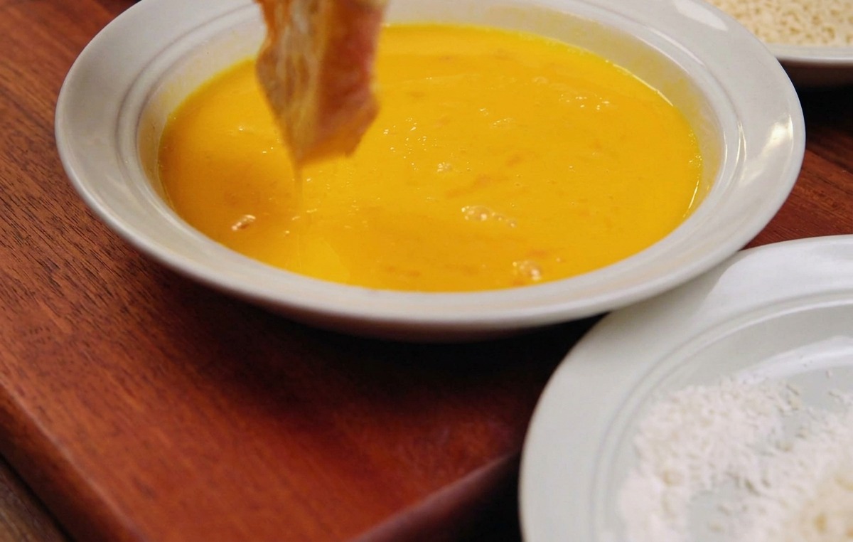 A slice of pork being dipped into a shallow white bowl filled with a bright yellow egg wash mixture.