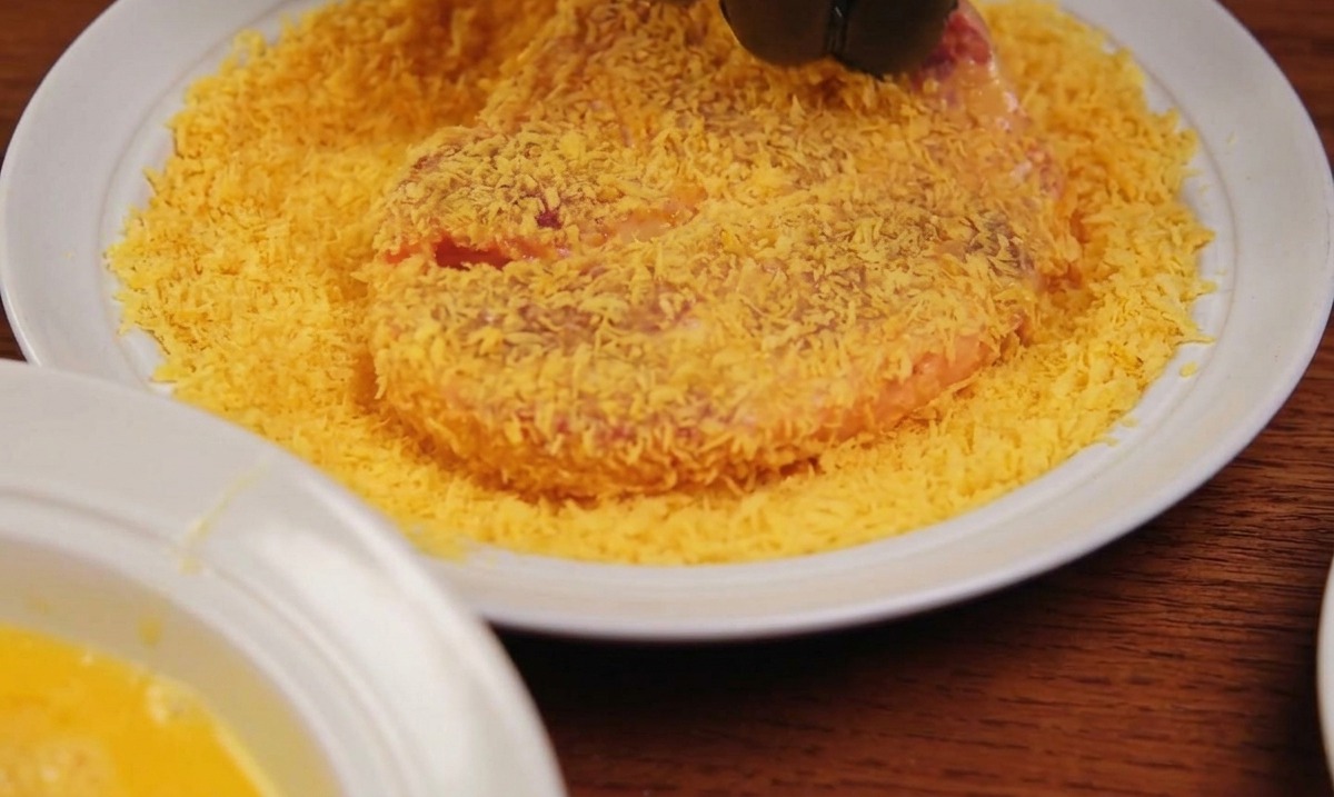 A hand in a black glove pressing a piece of meat into a plate covered in golden panko breadcrumbs.
