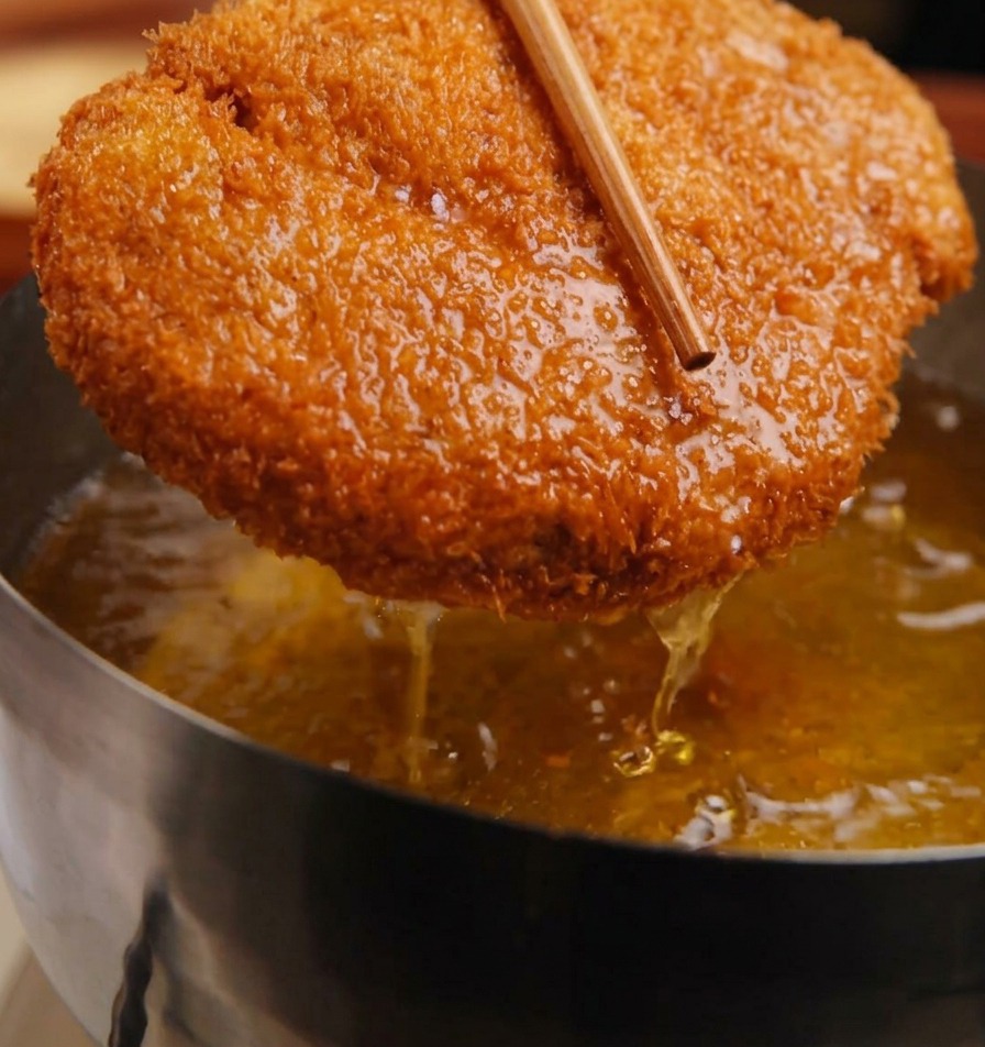 A golden, panko-crusted pork cutlet being held with wooden chopsticks above a pot of bubbling oil.