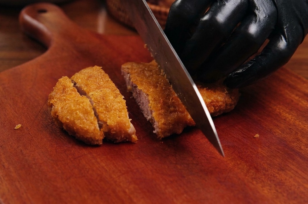 A sharp chefs knife slicing through a golden-brown, breaded fried pork cutlet on a wooden board.