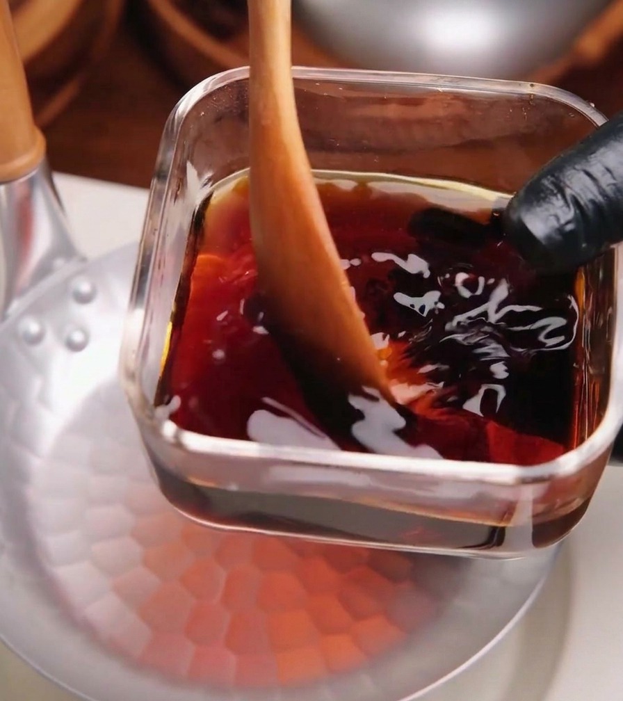 Dark katsudon sauce being poured from a glass container into a shallow, hammered metal cooking pan.