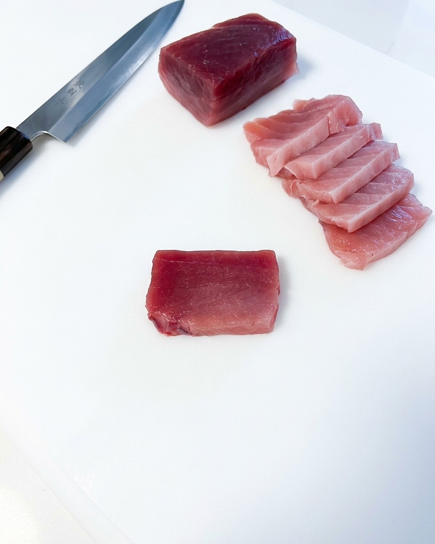Several rectangular slices of pink fatty tuna arranged next to a large block of tuna and a Japanese kitchen knife on a white board.