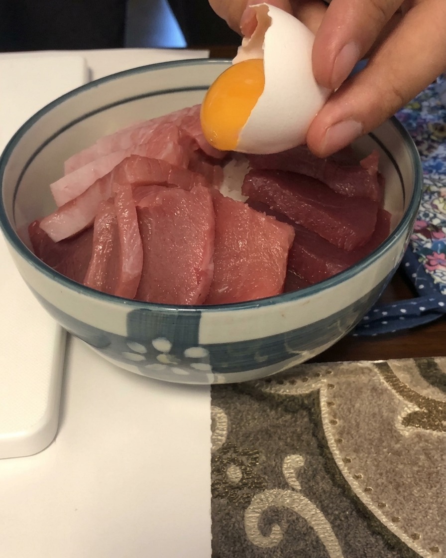 A bright yellow egg yolk sliding out of a white eggshell onto a bed of sliced raw fish in a blue and white bowl.