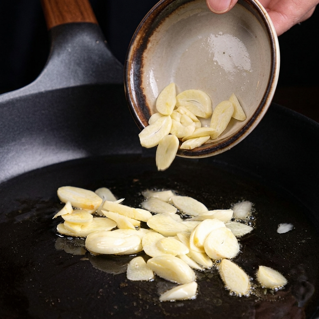 Sliding raw garlic slices from a small bowl into a hot pan with oil to fry.