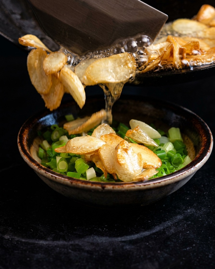 Pouring sizzling hot oil and fried garlic chips from a skillet into a bowl of chopped green onions.
