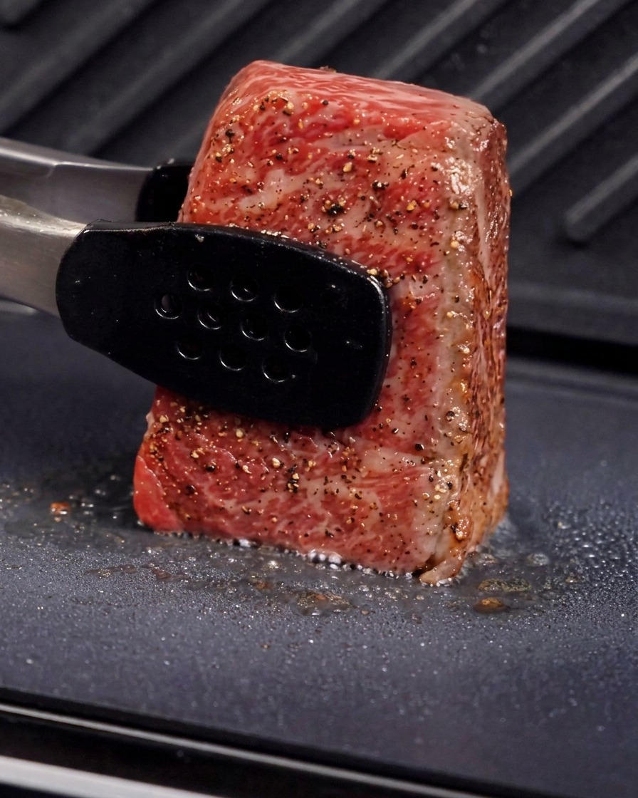 Metal tongs holding a thick piece of beef upright on a grill plate to sear the side, with visible steam and sizzling oil.