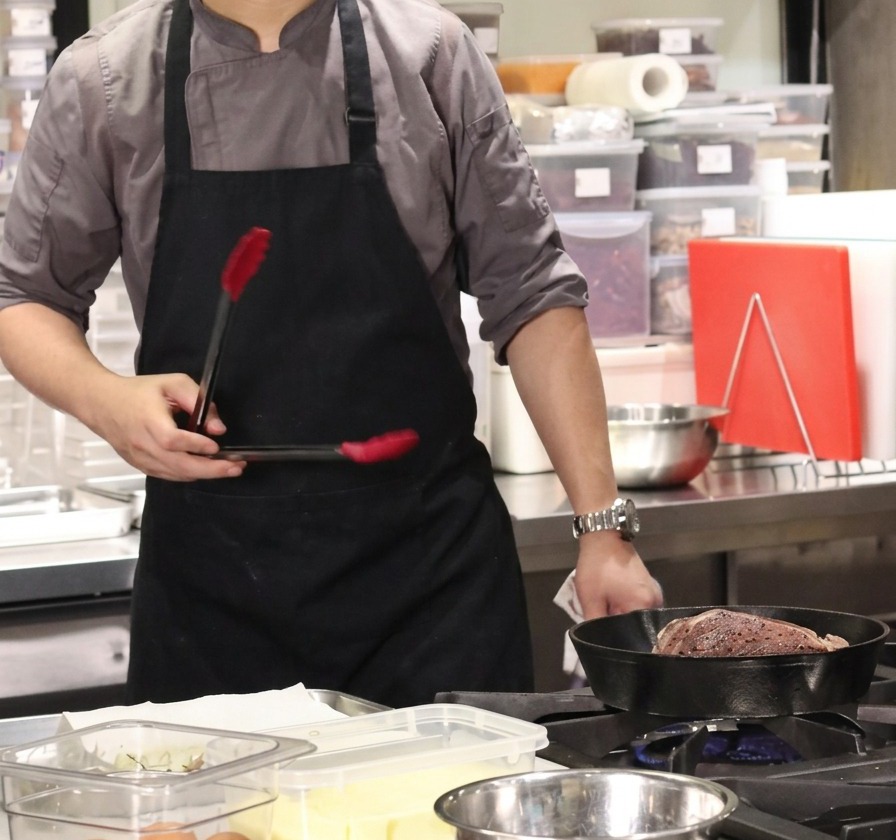 A chef holding tongs above a hot cast-iron skillet containing a large beef block to sear the fat layer.