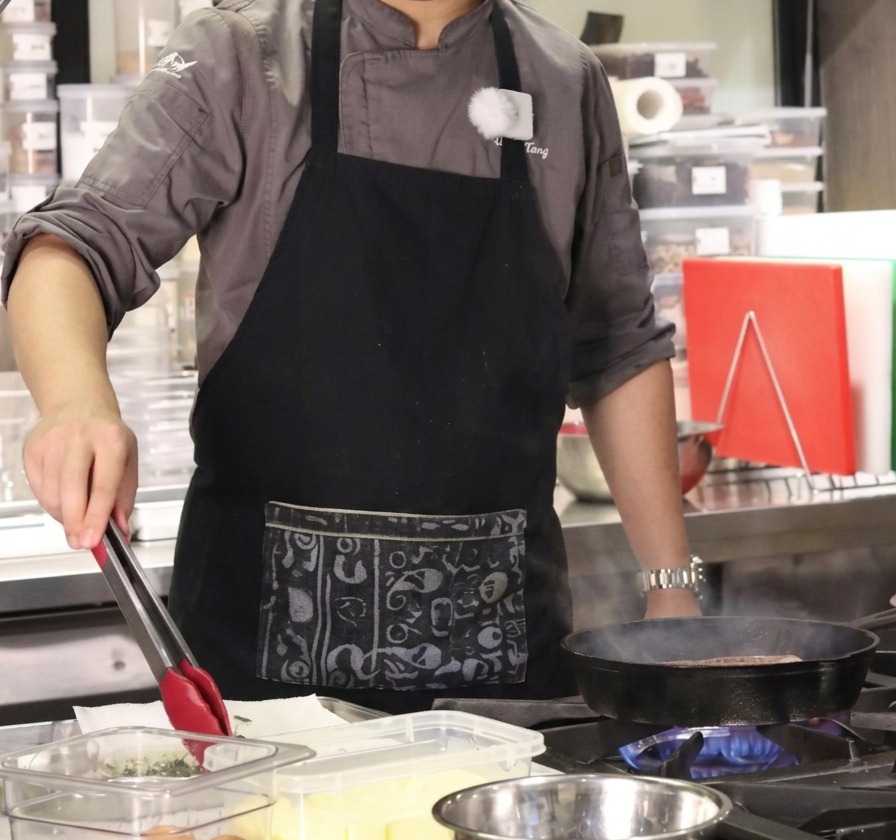The chef using tongs to transfer fresh thyme sprigs and garlic from a clear container into the pan.
