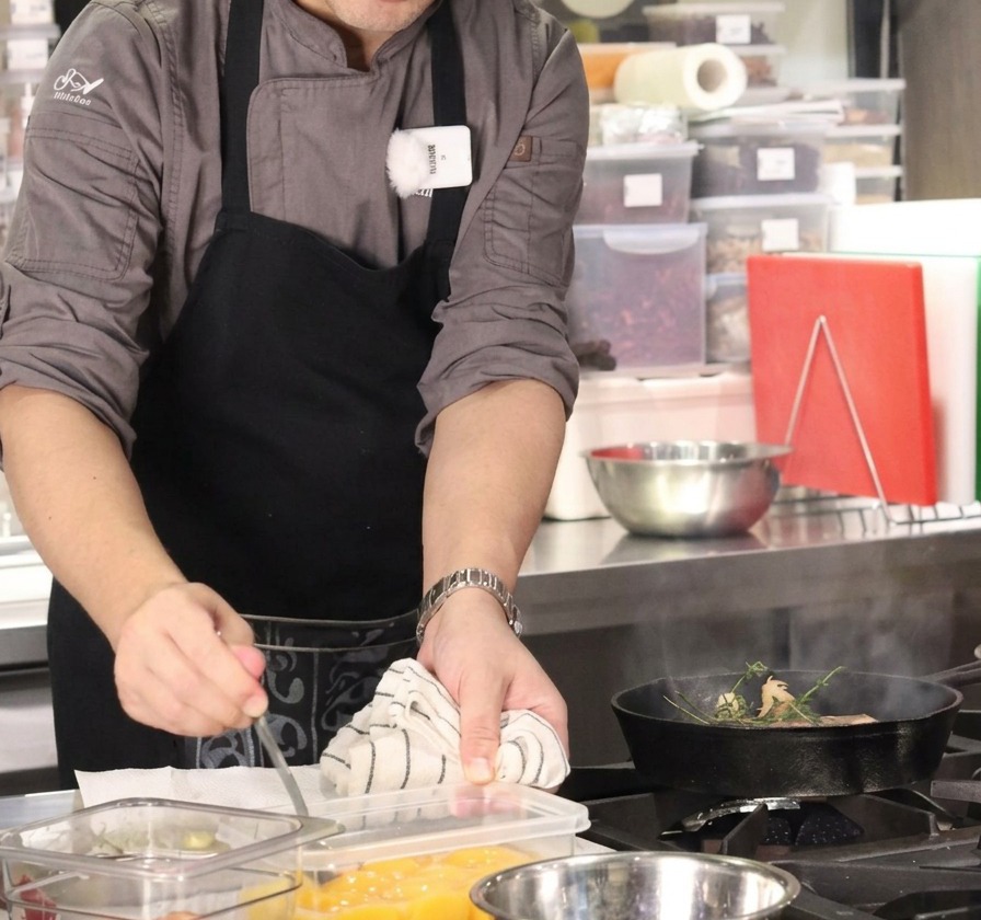 A chef using a spoon to add a cube of butter from a prep container into a smoking pan with beef and herbs.