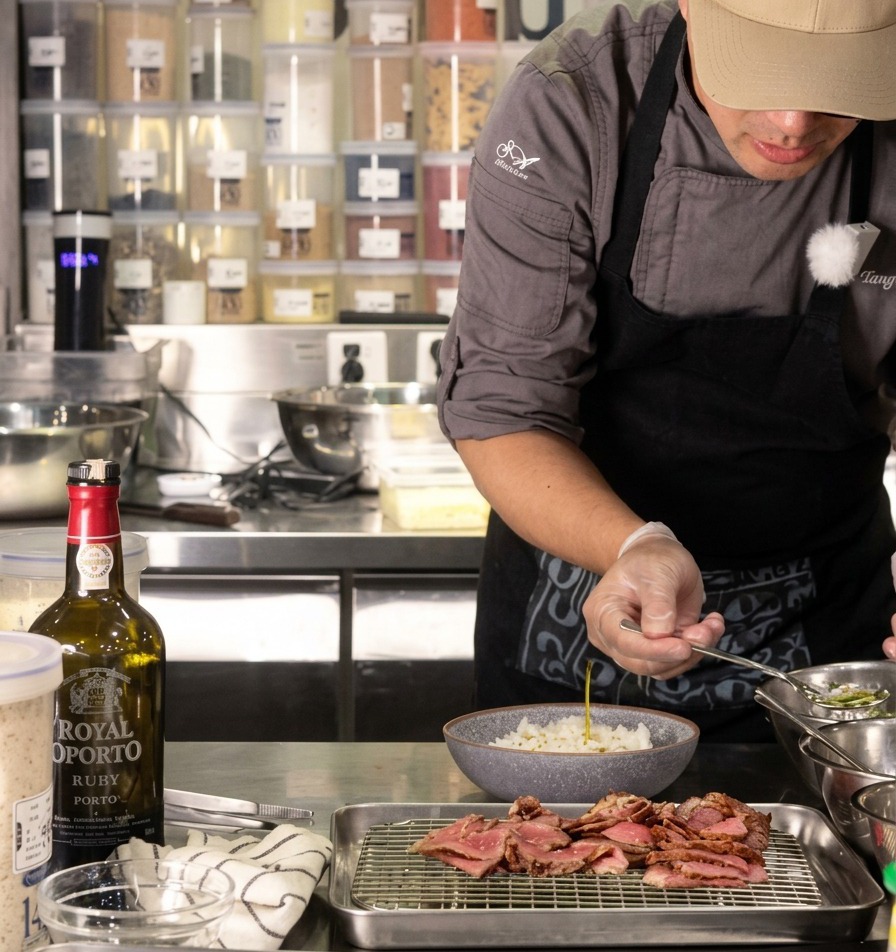 A chef pouring a small spoonful of clear scallion oil over a bowl of white rice, with sliced beef resting on a wire rack nearby.