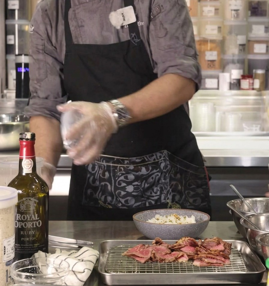 The chefs hand shaking savory furikake seasoning onto a bowl of rice on a stainless steel kitchen counter.
