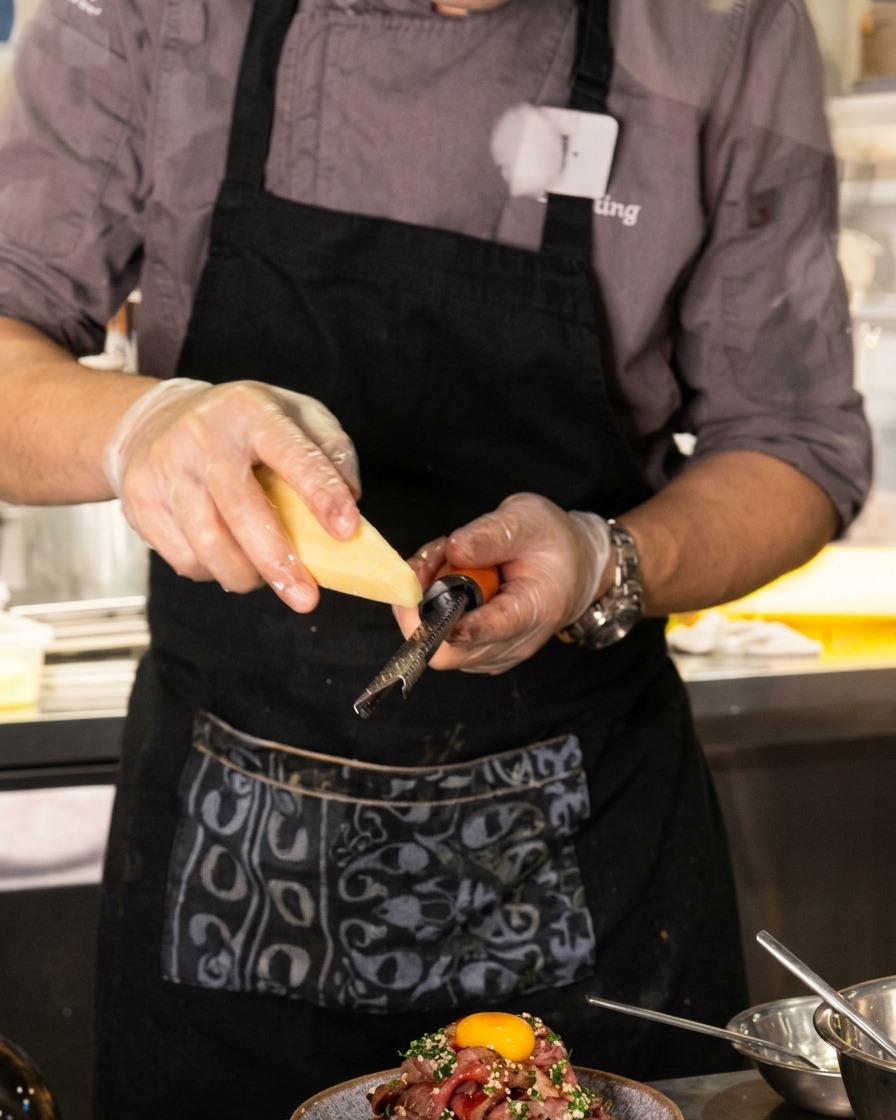 A close-up shot of a chef using a microplane to finely grate cheese over a towering mountain of sliced beef topped with an egg yolk.