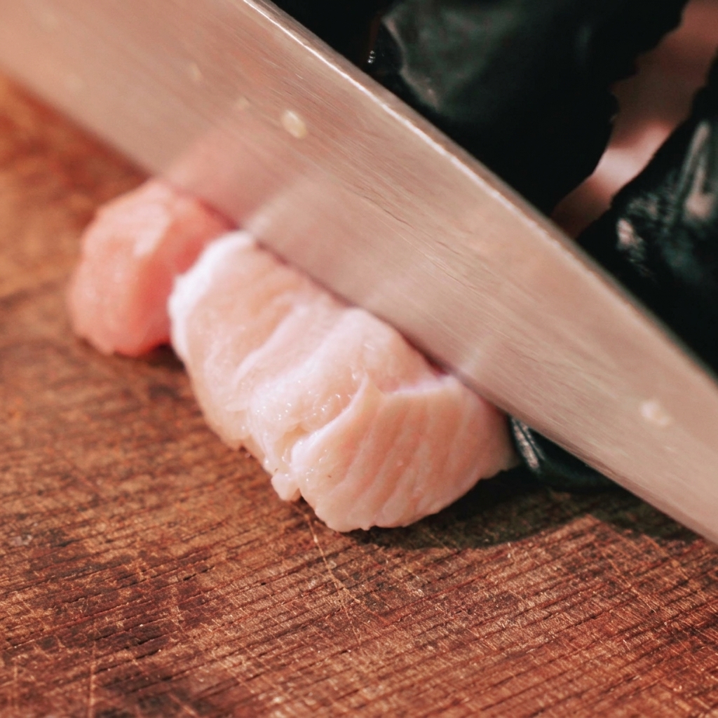 A gloved hand using a chefs knife to slice raw, skin-on pork belly into thick strips on a wooden cutting board.