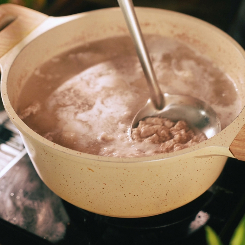 A metal ladle scooping grey foam and impurities from the surface of boiling water in a beige pot containing pork strips.