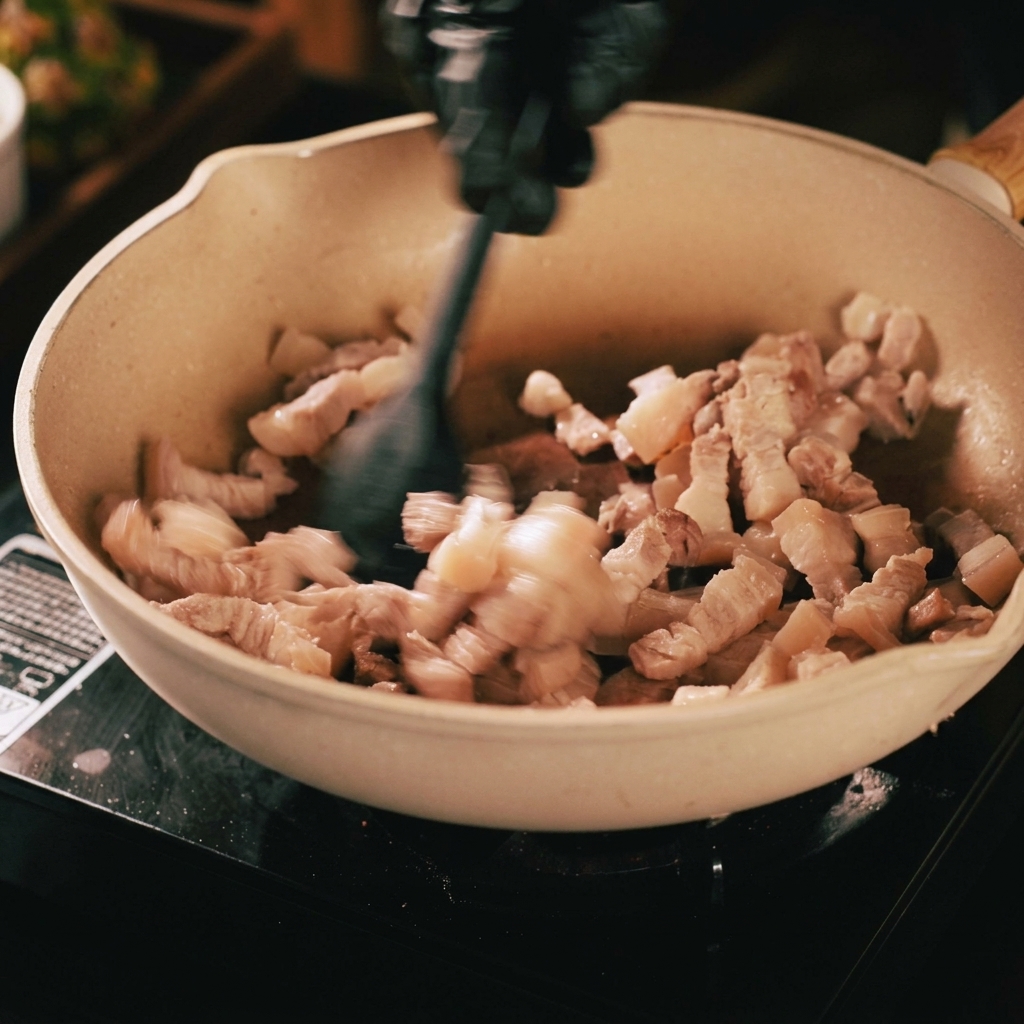Pork strips being stir-fried in a beige pan with a black spatula, showing browning and rendered oil.