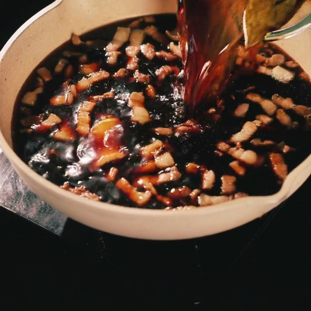 A dark savory marinade being poured from a glass container onto browned pork strips in a cooking pan.