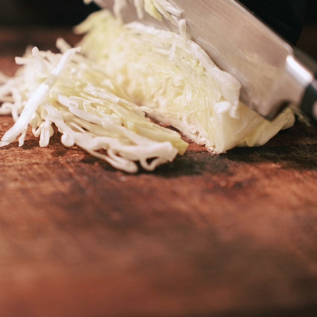 A close-up of a knife finely slicing through fresh, pale green cabbage on a wooden cutting board.