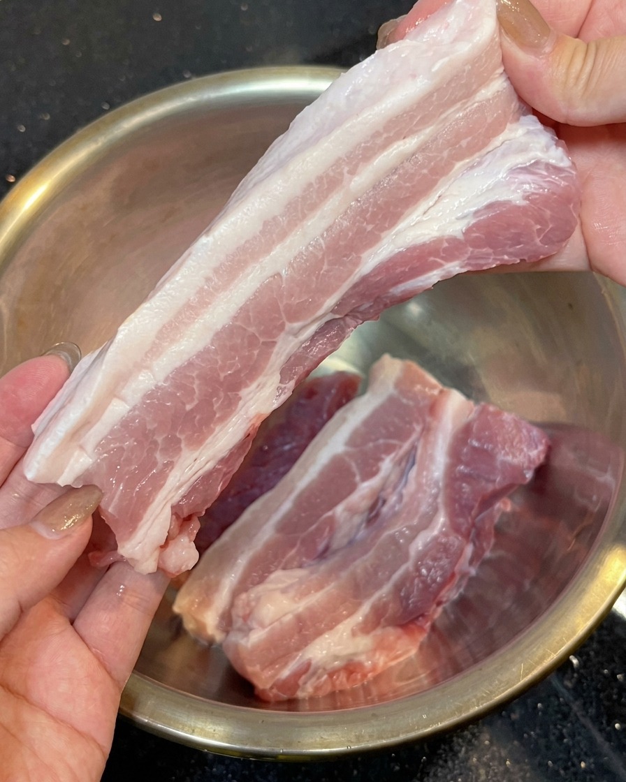 Two hands holding a long strip of pink and white layered raw pork belly with the skin side clearly removed, over a stainless steel bowl.
