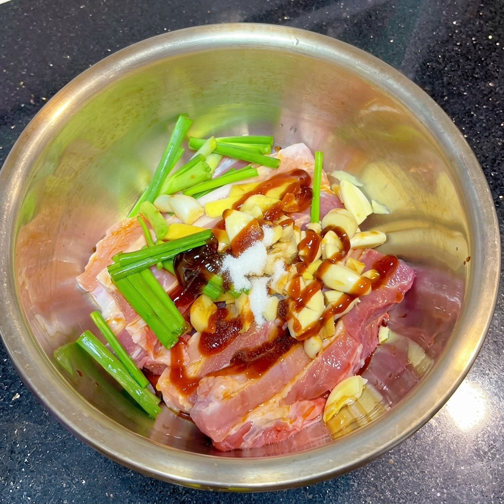 A pile of white salt and reddish-brown dry spice powder sitting on top of marinated pork strips and aromatics in a metal bowl.