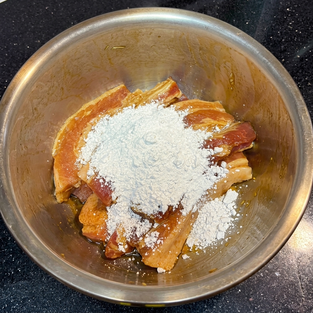 A large mound of white, powdery sweet potato starch sitting on top of dark brown marinated pork strips in a bowl.
