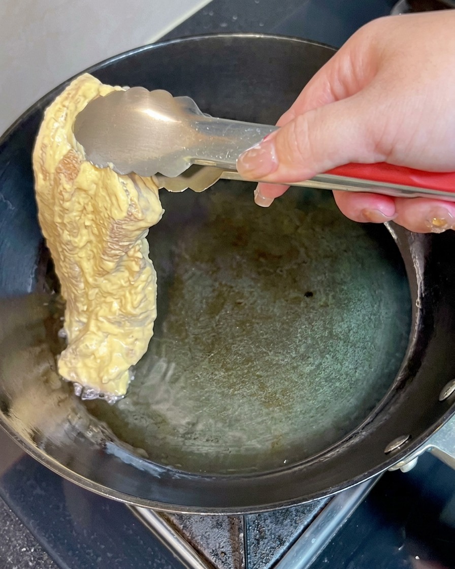A piece of battered pork belly being gently lowered into hot oil in a pan using metal tongs.