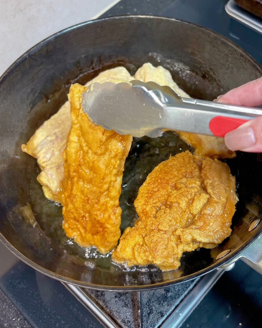 Metal tongs flipping a piece of crispy, golden-brown pork belly in bubbling hot oil alongside other pieces.