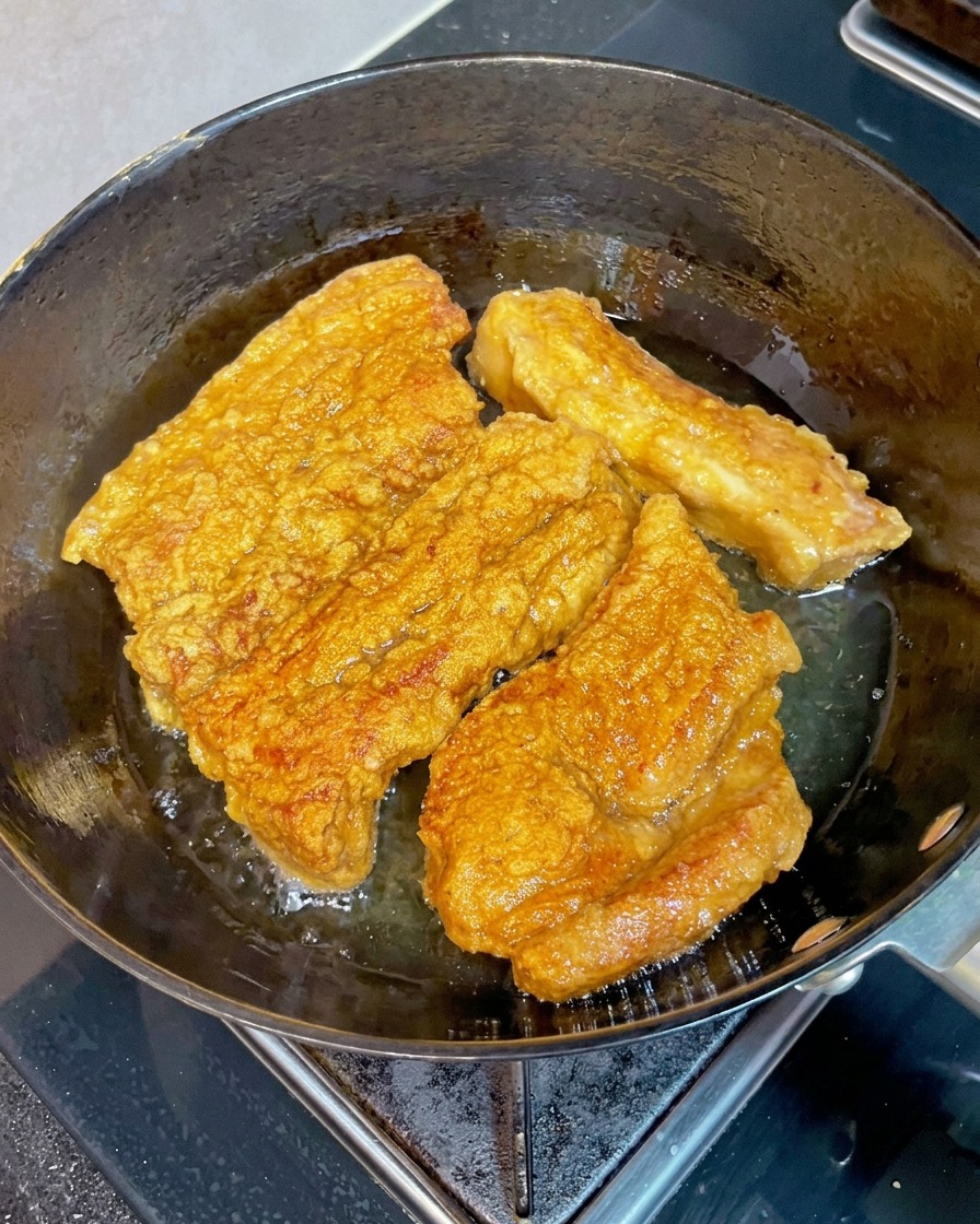 Crispy golden brown slabs of pork belly frying in a pan of hot oil with a wooden chopstick pressing into one piece to check if it is tender.