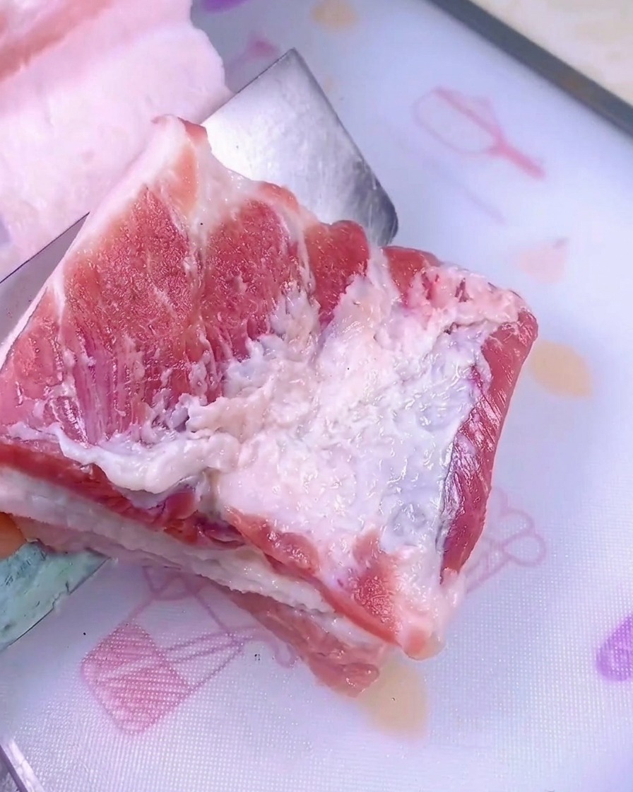 A sharp knife slicing through a thick slab of raw pork belly on a white plastic cutting board.