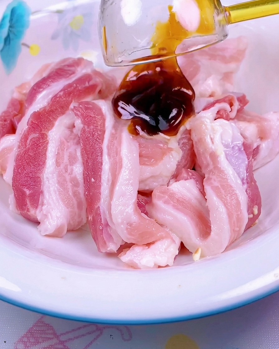 Dark red sauce being drizzled over a pile of raw, sliced pork belly in a white ceramic bowl.